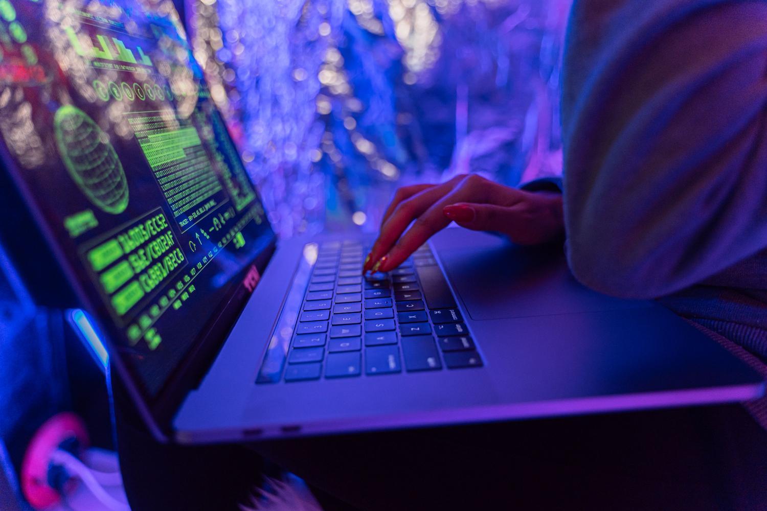 Ciberseguridad en portátil Manos escribiendo en un ordenador portátil con gráficos de ciberseguridad en pantalla y luces ambientales azules y púrpuras.
