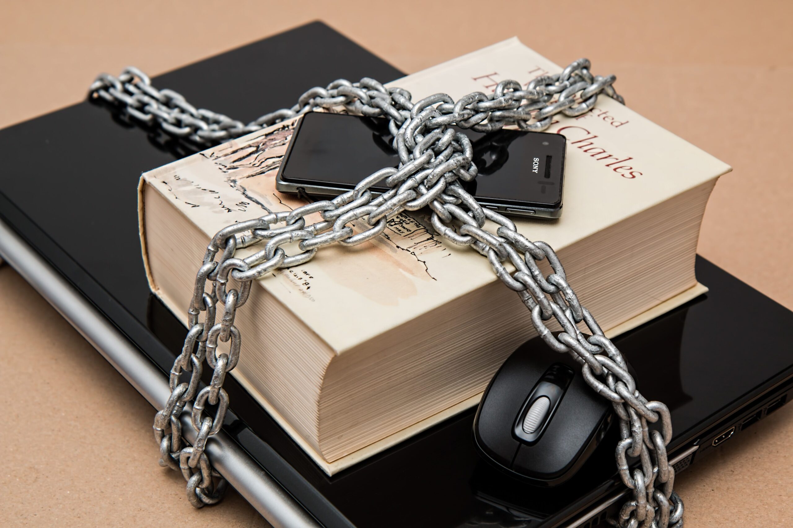 Dispositivos tecnológicos y un libro encadenados con un candado, simbolizando las restricciones del software privativo y las licencias cerradas. Dispositivos tecnológicos y un libro encadenados con un candado, simbolizando las restricciones del software privativo y las licencias cerradas.