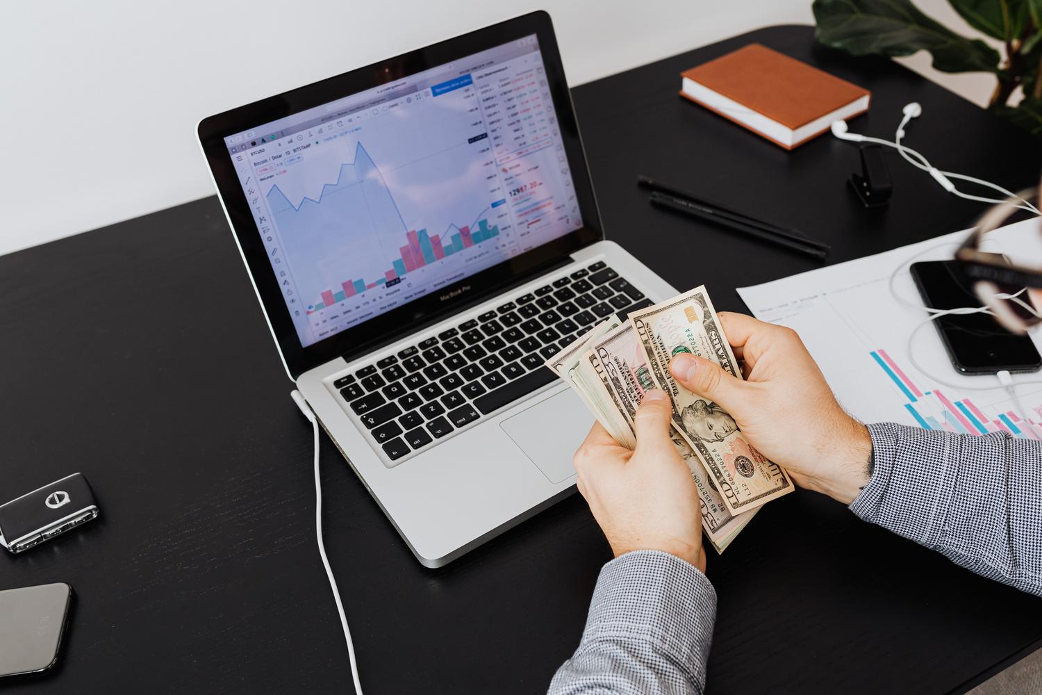 Persona contando billetes de dólar con gráficos financieros en una pantalla de portátil, representando la rentabilidad económica y el beneficio real. Persona contando billetes de dólar con gráficos financieros en una pantalla de portátil, representando la rentabilidad económica y el beneficio real.