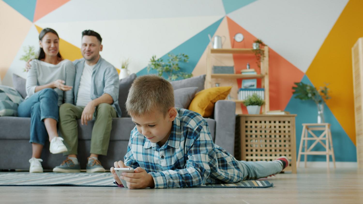 Control parental discreto Un niño utiliza un smartphone mientras sus padres lo observan atentamente desde el fondo, ilustrando la supervisión y el control parental discreto.