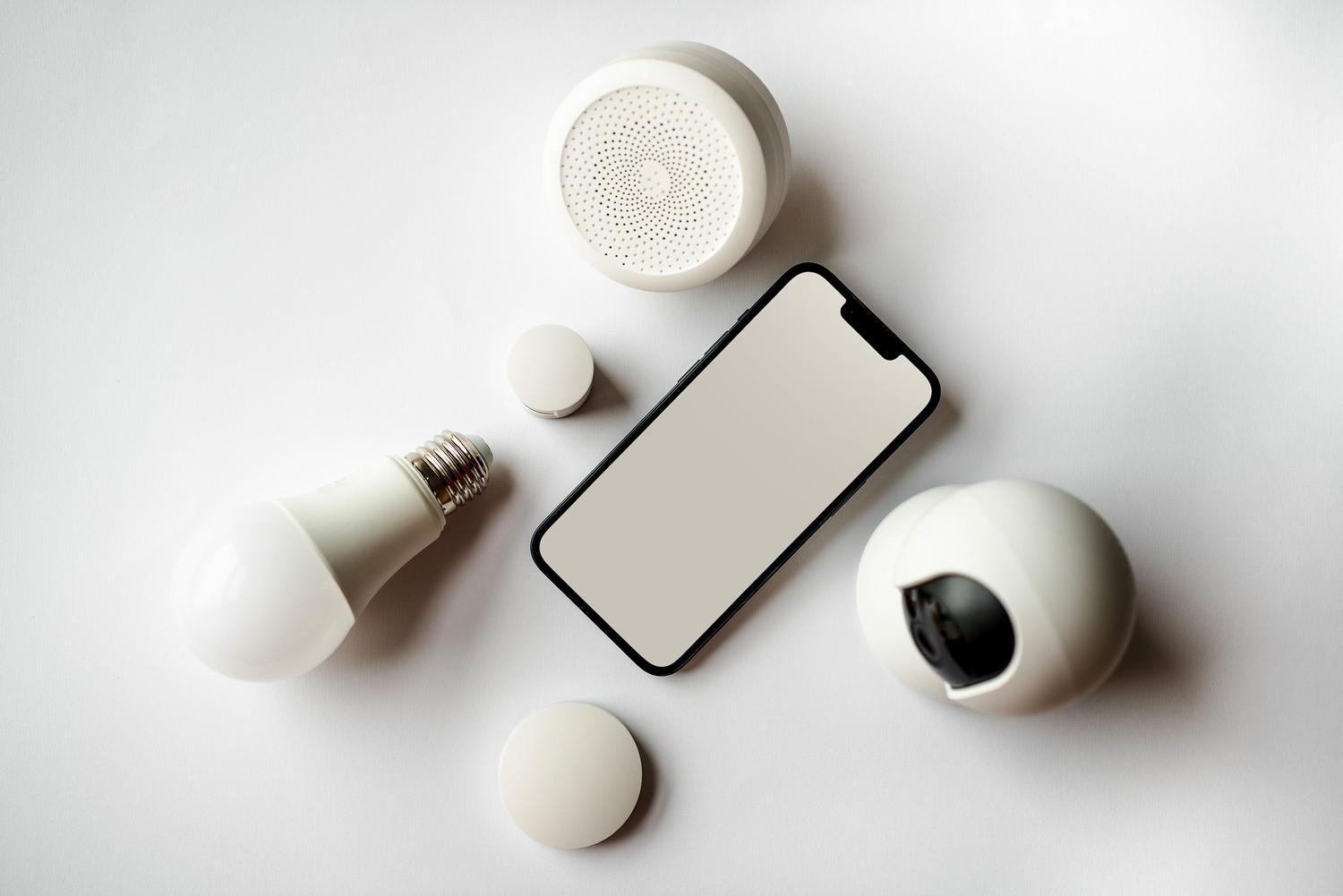 Vista cenital de un smartphone y diversos dispositivos del hogar inteligente sobre fondo blanco, mostrando la saturación potencial de conexiones. Vista cenital de un smartphone y diversos dispositivos del hogar inteligente sobre fondo blanco, mostrando la saturación potencial de conexiones.