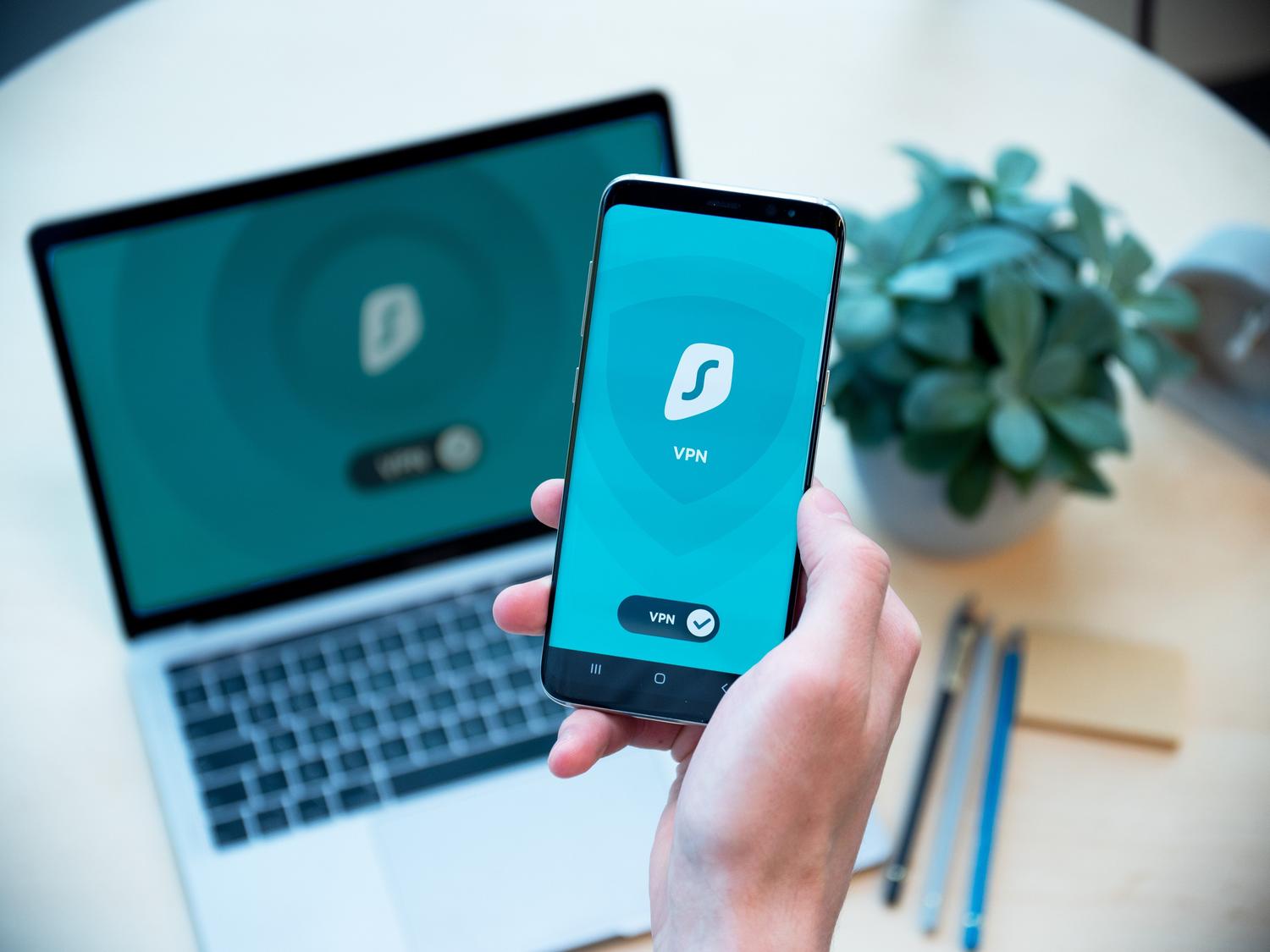 Mano sosteniendo un smartphone con un escudo de seguridad activo y un portátil al fondo, representando la prevención y seguridad digital. Mano sosteniendo un smartphone con un escudo de seguridad activo y un portátil al fondo, representando la prevención y seguridad digital.