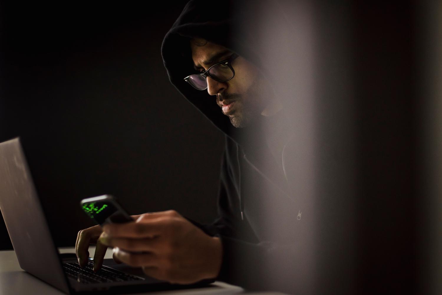 Un ciberdelincuente en una habitación oscura con capucha y gafas, iluminado por la luz de un portátil y un móvil, representando el robo de cuentas digitales. Un ciberdelincuente en una habitación oscura con capucha y gafas, iluminado por la luz de un portátil y un móvil, representando el robo de cuentas digitales.